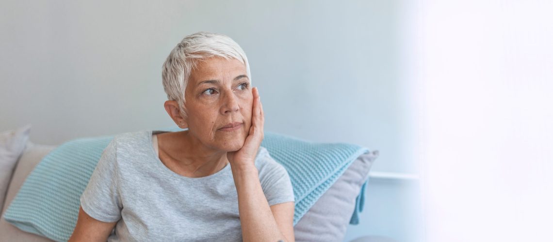 Upset senior woman looking in distance remembering old times, sad aged female sit at home feeling lonely missing passed husband, elderly lady crying near window having difficult life situation