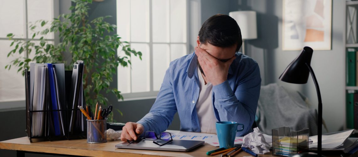 Close up of young man sitting at workplace at home office. Tired businessman rubbing eyes, feeling exhausted. Daily routine.